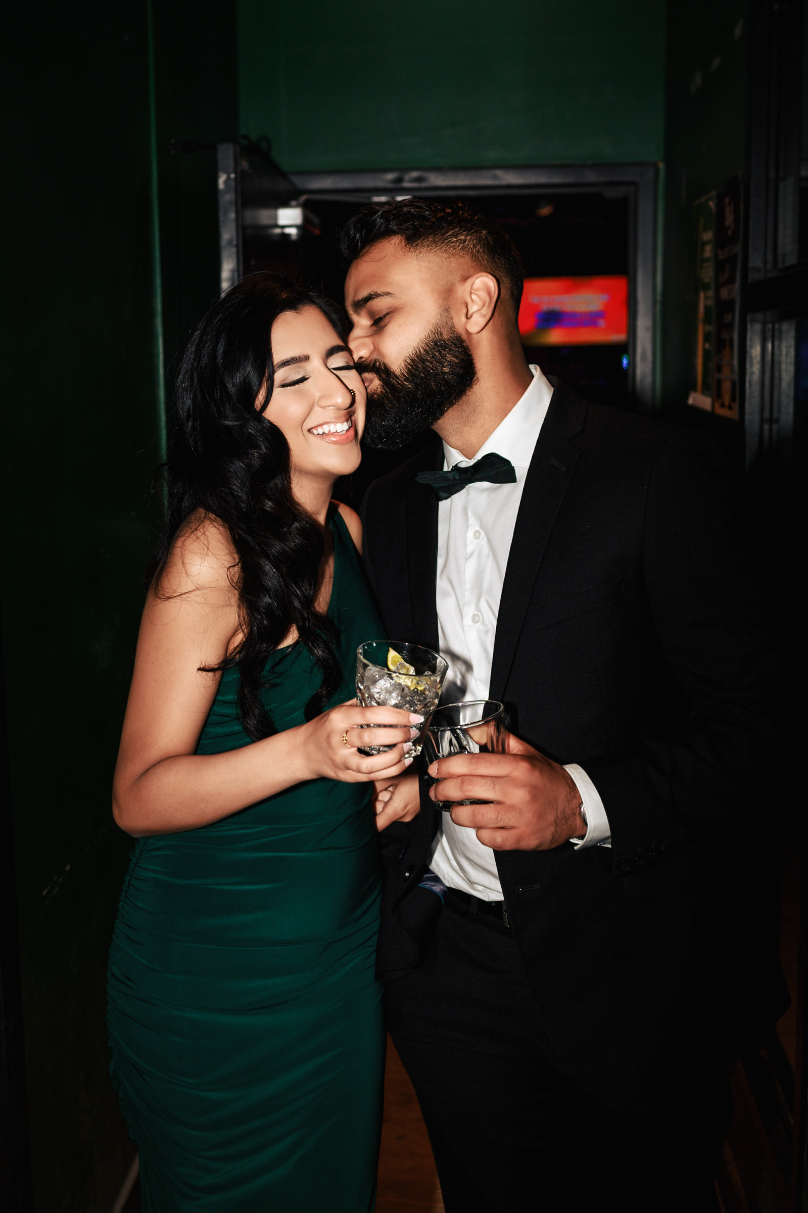 A couple dressed in formal attire shares a tender moment during their Indian engagement shoot. The woman in a green dress holds a drink while smiling with her eyes closed. The man in a black suit with a bow tie kisses her cheek and holds his own drink, creating a warm and intimate scene.