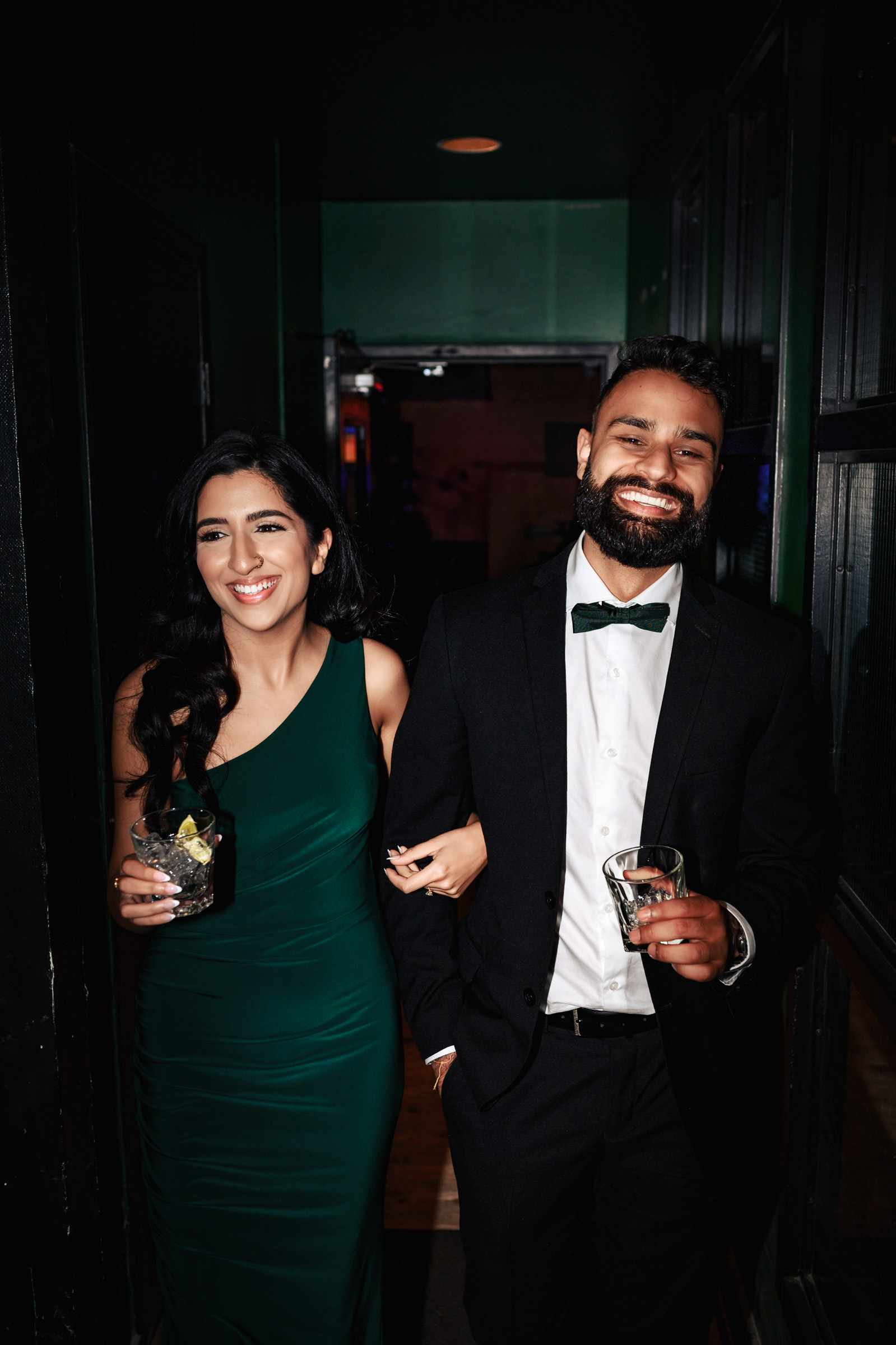 A woman in an elegant green dress and a man in a black suit with a green bow tie smile and walk arm-in-arm, each holding a drink in their other hand. They appear to be at an indoor Indian engagement shoot, with dim lighting enhancing the celebratory atmosphere.