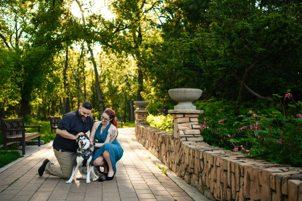 A man and woman kneel on a brick pathway, smiling as they pet a joyful dog wearing a harness. They are surrounded by greenery and flowers, with a stone wall and garden urns in the background. It is a bright, sunny day in what appears to be a park.