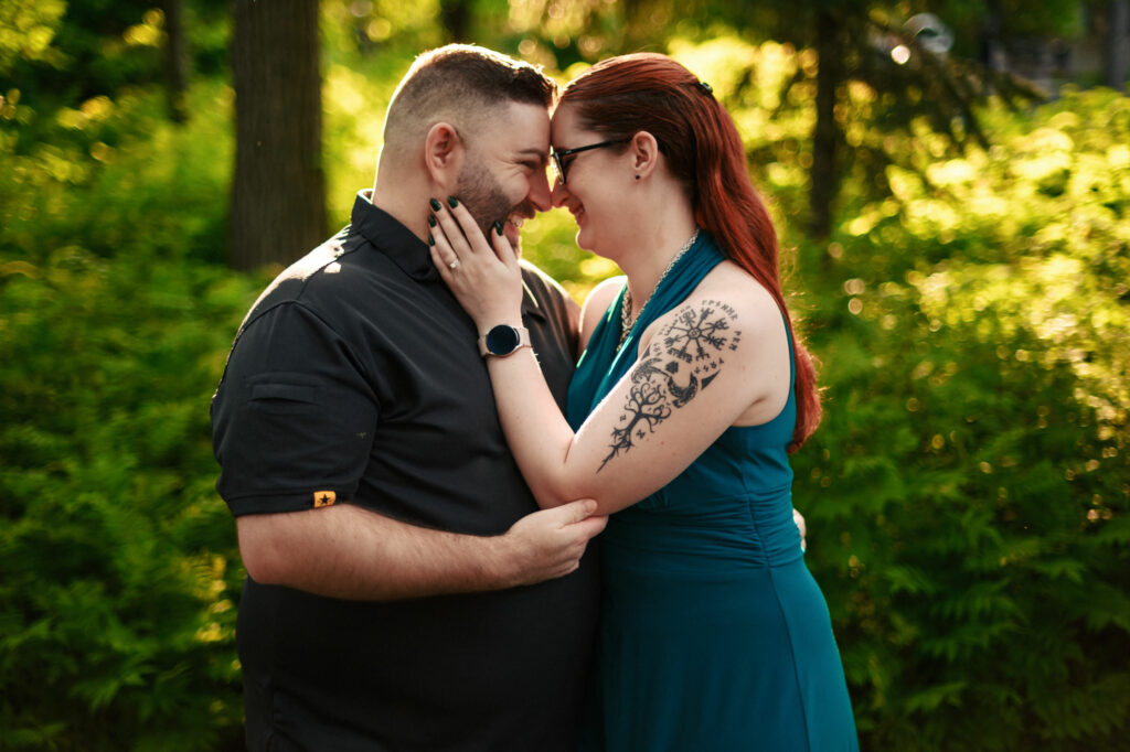 A couple stands close together outdoors, embracing and touching foreheads. The man wears a dark shirt, and the woman wears a teal dress and glasses, with one hand on his cheek, revealing a floral tattoo on her arm. The background is lush and green, lit by sunlight.