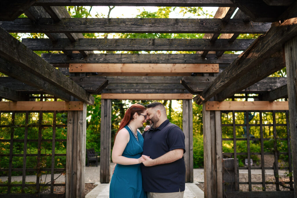 A couple stands under a wooden pergola surrounded by greenery. They are facing each other, holding hands, and smiling. The woman with red hair wears a teal dress, while the man with facial hair wears a black shirt. The sunlight filters through the structure above them.