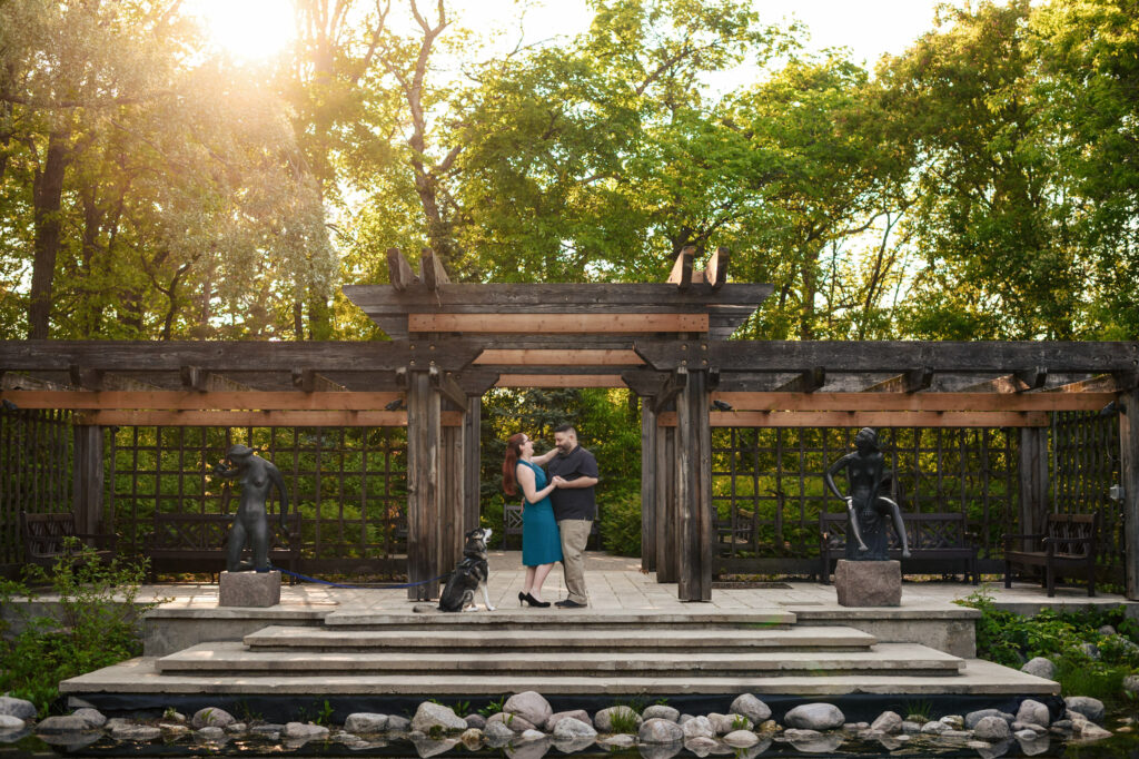 A couple embraces under a wooden pergola in a garden with lush green trees. The area is adorned with sculptures and surrounded by serene nature. Stones are scattered near a water feature in the foreground, adding to the tranquil ambiance.