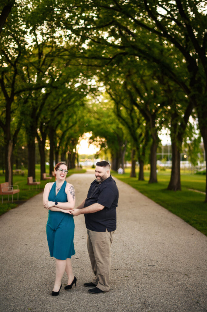 A couple stands on a tree-lined pathway at sunset. The woman, wearing a teal dress and glasses, smiles while the man in a black shirt and khaki pants gently holds her forearm. Both seem happy, sharing a light moment against the backdrop of lush greenery.