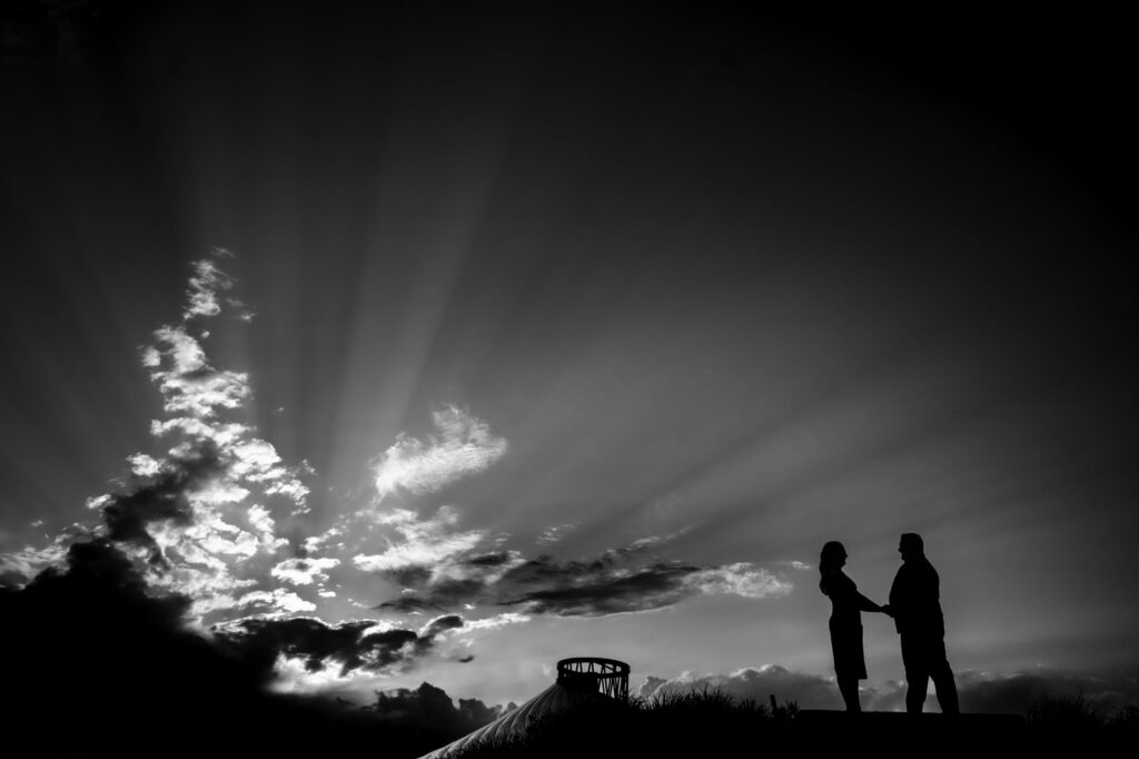 A black-and-white silhouette of two people holding hands, standing on a hill with a cloudy sky and rays of sunlight breaking through. A circular structure is visible in the background.