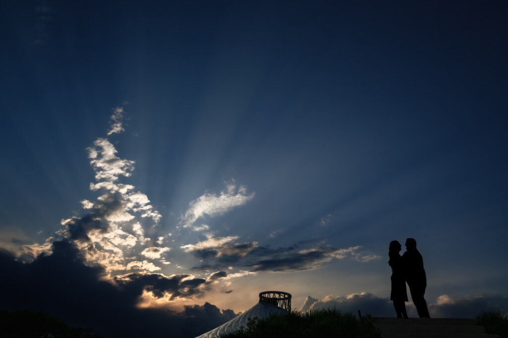 Silhouetted couple stand close together, facing each other against a dramatic sky with rays of sunlight streaming through scattered clouds at sunset. Below, greenery and a curved metallic structure are partially visible.