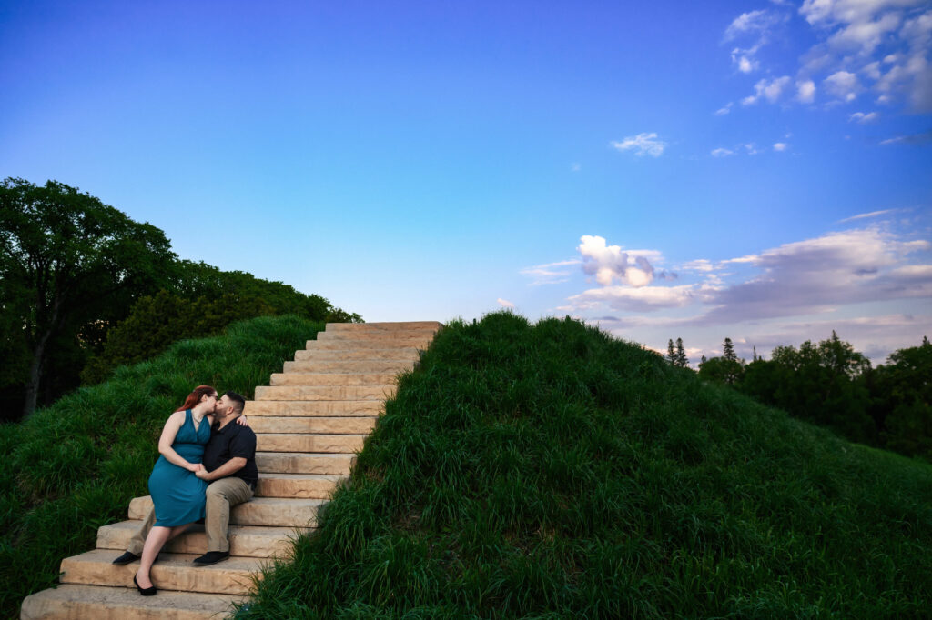 A couple sits together on stone steps leading up a grassy hill, kissing. The sky is clear with a few clouds, and trees are visible in the background. They appear to be enjoying a peaceful moment outdoors during sunset.