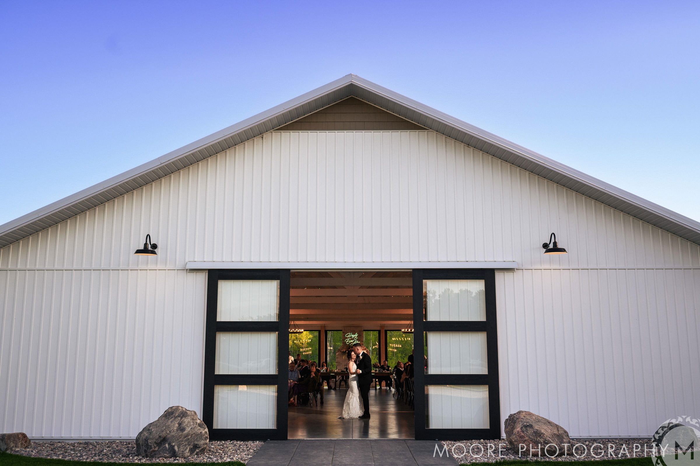 white poplar wedding A couple shares their first dance inside a large, white barn with modern black sliding doors. The barn is surrounded by stones, and warm lights illuminate the scene at dusk. The barn's interior is visible, with guests seated at tables in the background.