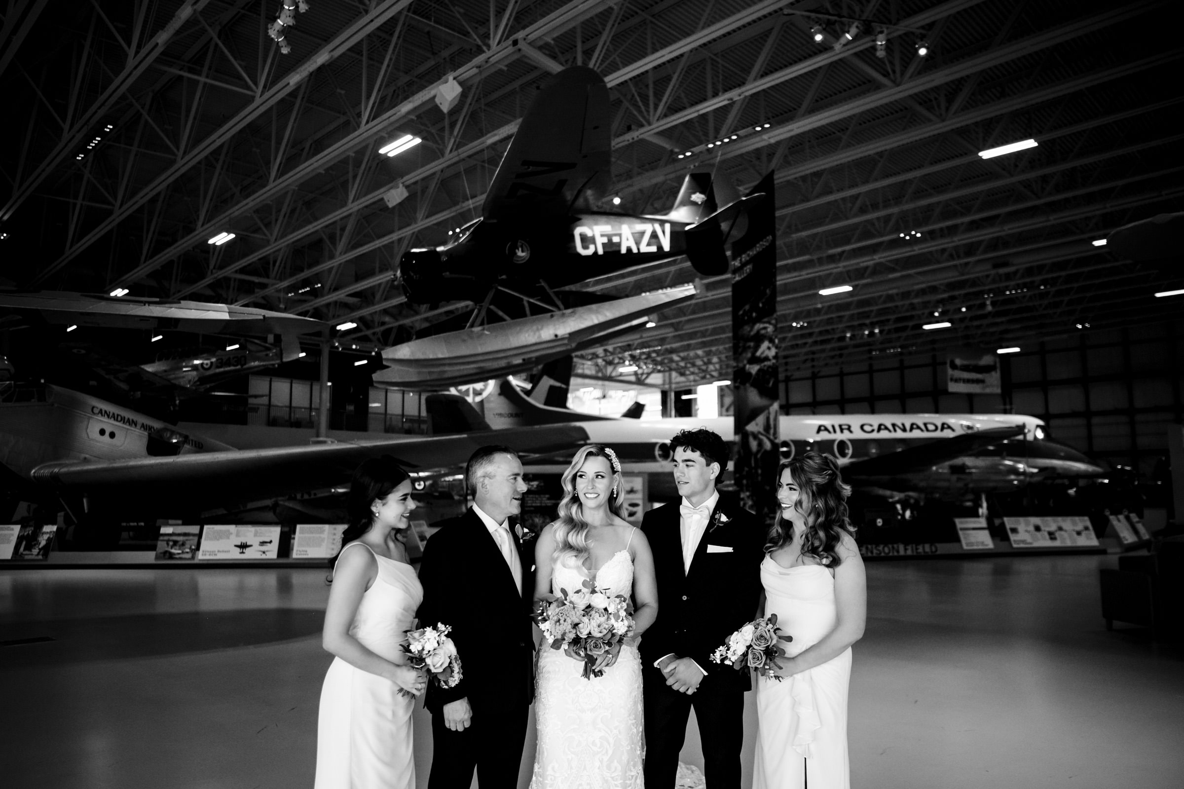 Winnipeg wedding party posing by vintage aircraft in a hangar, captured in black-and-white.