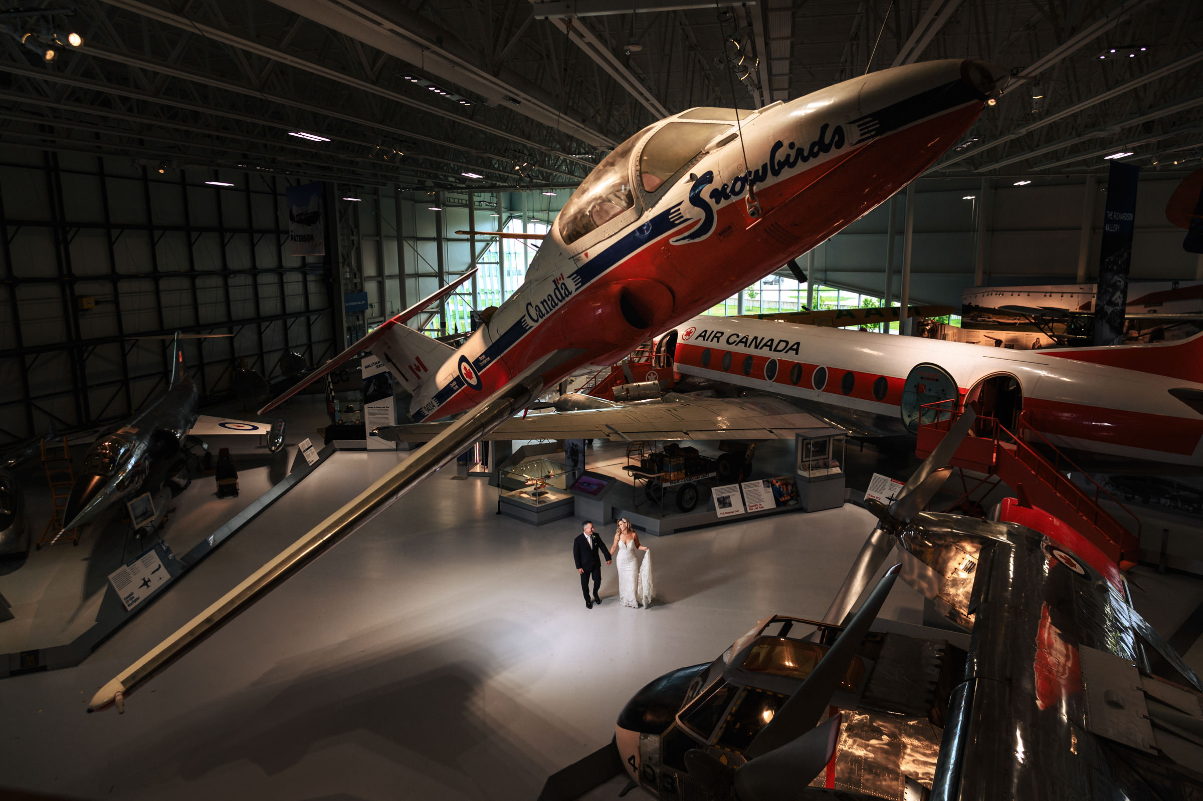 Bride and groom walk in a Winnipeg wedding venue under a large aircraft display.