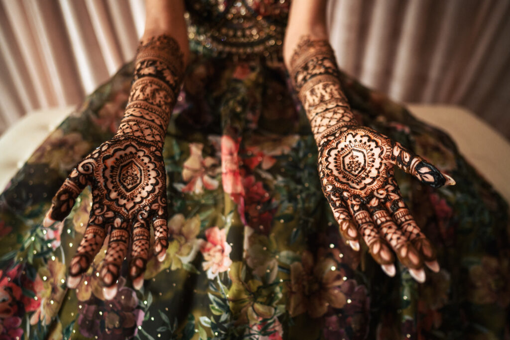 A person in a floral dress displays intricate henna designs on their palms and forearms. The henna patterns feature elaborate motifs and geometric shapes, showcasing traditional artistry. The background is softly blurred, emphasizing the focus on the henna.