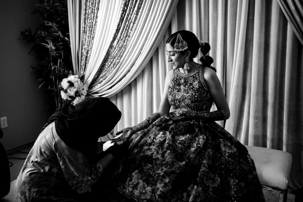 A woman seated in a flowing, intricately designed gown, possibly a wedding dress, has her hands adorned with henna designs by another person. The background features ornate curtains and floral decorations. The scene suggests a traditional ceremonial preparation.