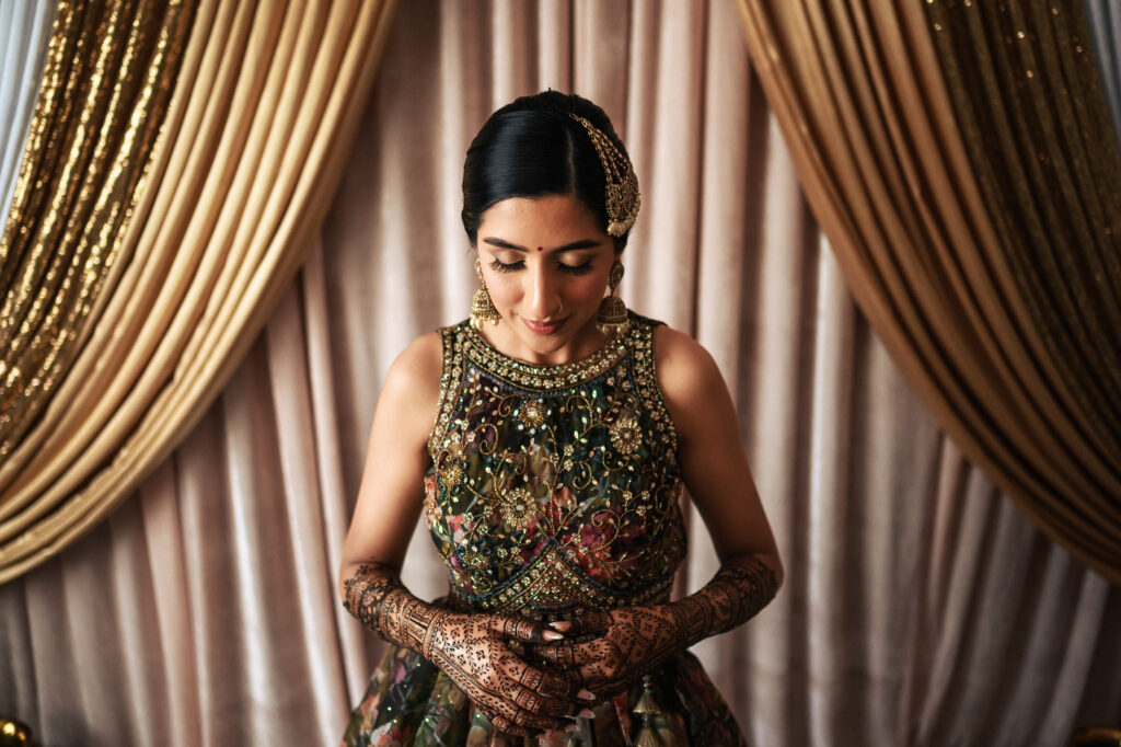 A woman wearing intricate floral attire stands with her eyes closed and hands clasped, displaying detailed henna designs on her arms. She is adorned with traditional jewelry and stands against a backdrop of golden curtains.