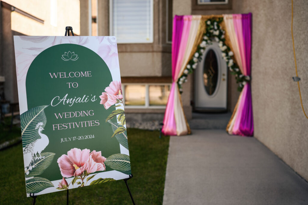 A sign reads, "Welcome to Anjali's Wedding Festivities, July 17-20, 2024," decorated with floral and leafy graphics. In the background, a doorway is adorned with pink and white drapes, and a flower garland. The setting appears to be outdoors.