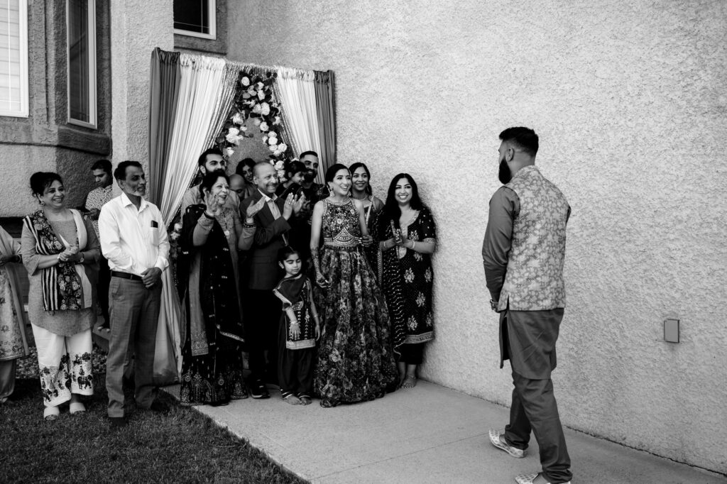 A group of people dressed in traditional South Asian attire stand outside a building, smiling and clapping. A woman, possibly the bride, wearing an ornate dress, stands at the center with others around her. A man walks towards the group, likely the groom.