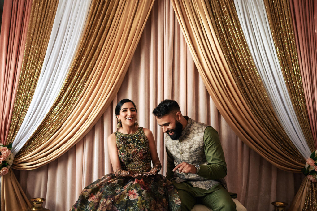 A couple dressed in traditional attire sits together, laughing. In the background, elegant drapes in shades of gold, pink, and white create a luxurious setting. The woman wears a detailed, ornate dress, while the man sports a patterned vest and green shirt.