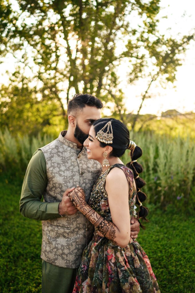 A couple is standing outdoors in a lush green field with trees in the background. The man is wearing a green shirt and a patterned vest, while the woman is in a colorful traditional dress with intricate jewelry and henna on her arms. They are smiling and holding hands.