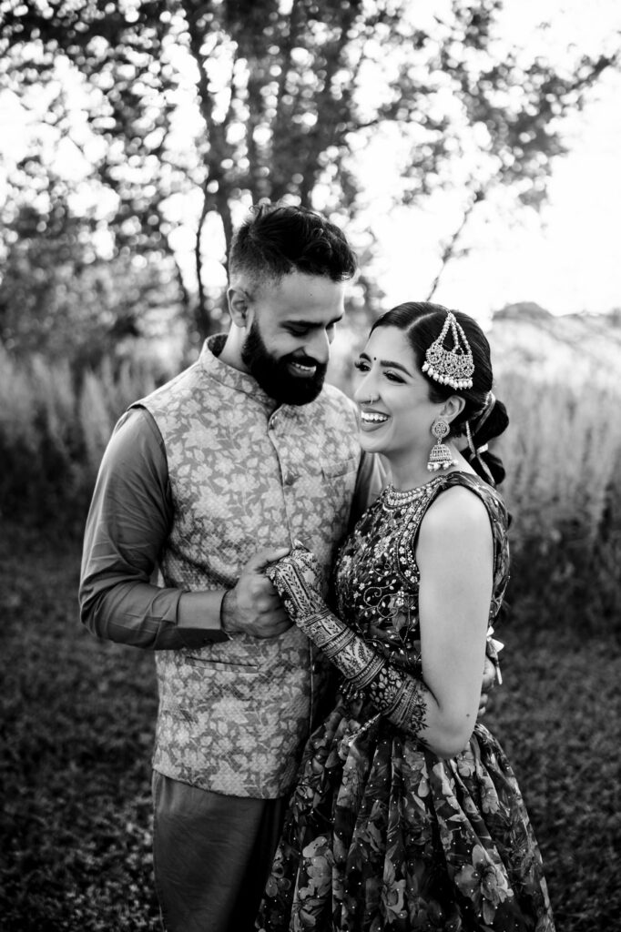 A couple joyfully holding hands, dressed in traditional attire, stands outdoors with trees in the background. The woman is wearing ornate, embroidered garments, and the man is in a patterned vest over a shirt. Both are smiling and looking at each other.