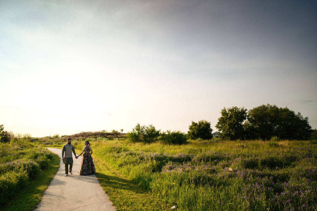 A couple walks hand-in-hand along a path through a sunlit meadow filled with grass and purple flowers. The sky above is clear and blue, with the warmth of the setting or rising sun casting a gentle glow over the scene. Trees line the horizon in the background.