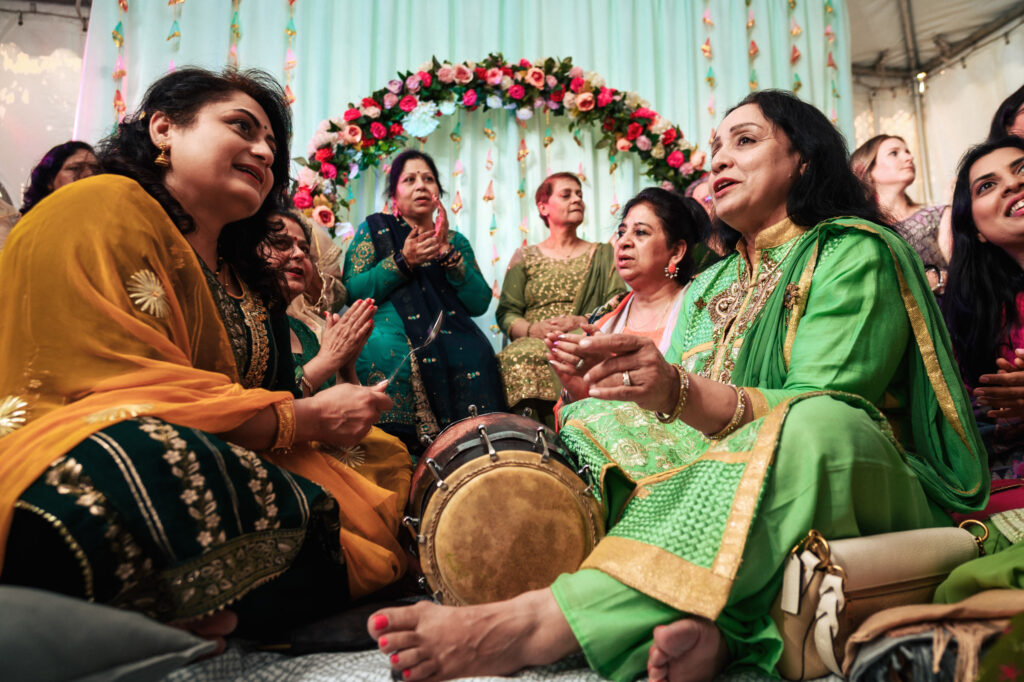 A group of women is sitting on the floor, dressed in colorful traditional attire, including green and yellow outfits. They are engaged in conversation and clapping, with one woman playing a dholak drum. Behind them is a floral arch and a light blue curtain.