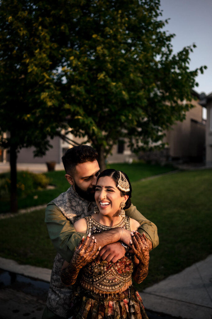A couple is embracing outdoors near a grassy area. The man has his arms around the woman from behind, and they both look joyful. They are dressed in traditional South Asian attire with intricate designs, and the woman has henna on her hands. A tree and buildings are in the background.