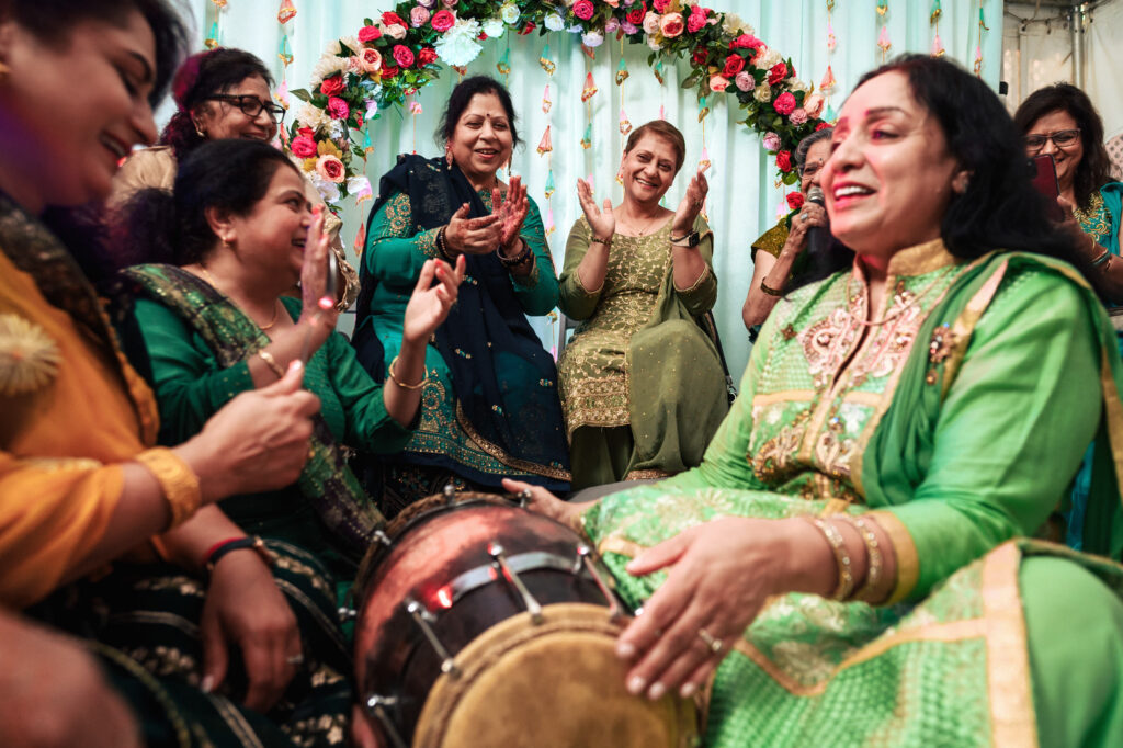 A group of women dressed in vibrant traditional attire joyfully sing and clap around a drum at a festive gathering. They are seated in front of a decorative backdrop adorned with a floral garland. Everyone is smiling and engaged in the celebration.