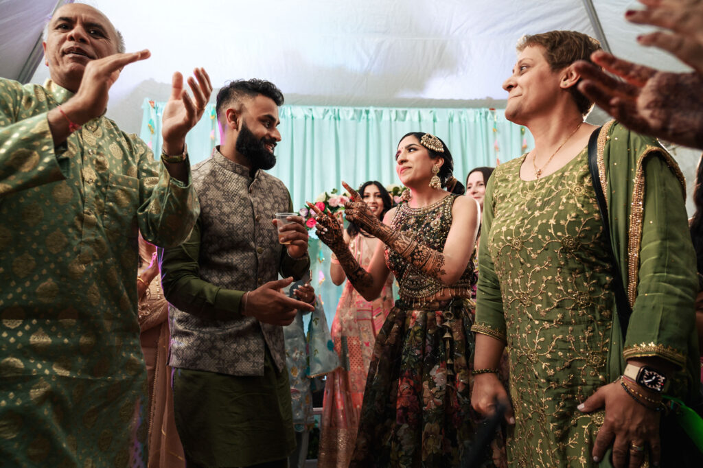 A joyful scene at a celebration shows a group of people in traditional attire. In the center, a man and woman share a lively moment under a tent. Others around them clap and smile, the atmosphere filled with happiness and festive decor.