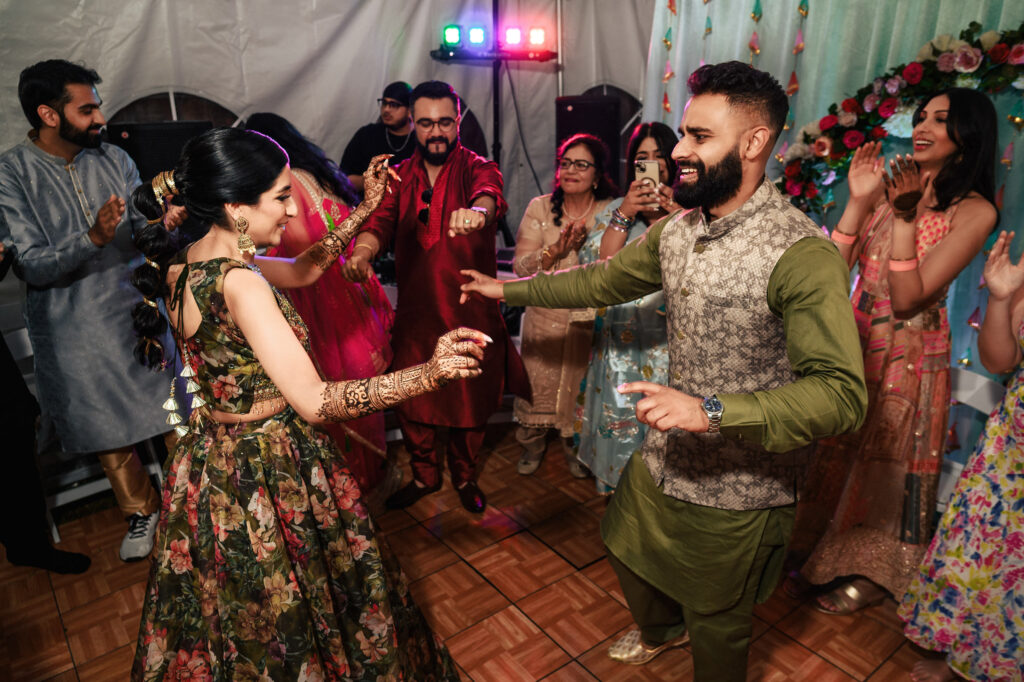 A lively celebration scene shows a group of people dancing joyfully. The participants are dressed in colorful traditional attire, with one woman in a floral dress and a man in a green outfit at the center. The background is decorated with flowers and lights.