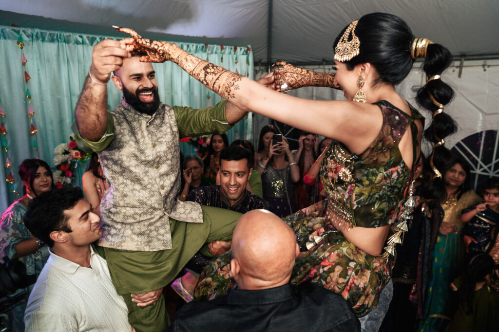 A joyful couple, dressed in traditional attire, is lifted by friends during a lively celebration. The bride and groom smile and reach out to each other, surrounded by guests who capture the moment on their phones. The festive atmosphere is vibrant and lively.