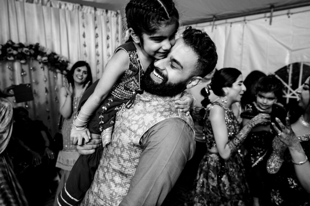 A black and white photo showing a man joyfully lifting a smiling young girl in his arms at a celebration. The girl is wearing traditional clothing with bangles, and other attendees in festive attire are seen in the background. The atmosphere is lively and cheerful.
