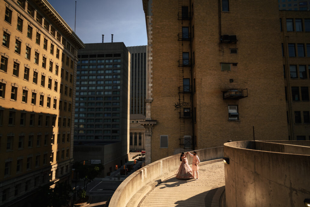 A couple dressed in wedding attire walks hand in hand on a curved outdoor pathway surrounded by tall buildings in a city. The bride is wearing a white dress and the groom is in a dark suit. The sun casts long shadows, adding a warm glow to the scene.