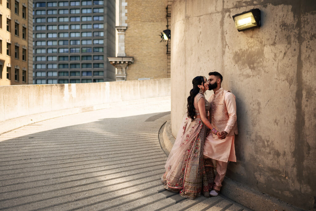 A couple dressed in traditional South Asian attire shares an intimate moment in an urban setting. The woman wears a peach-colored embellished lehenga, while the man wears a matching kurta-pajama. They stand close, holding hands, with a concrete wall and city buildings in the background.