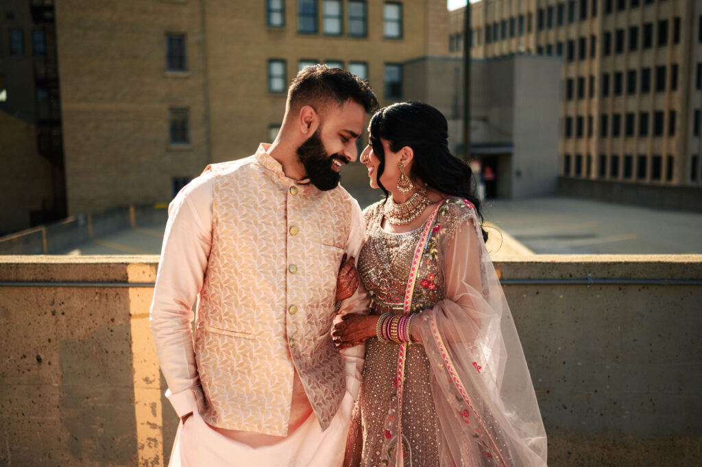 A couple dressed in traditional South Asian attire stands on an urban rooftop. The man in a light-colored sherwani smiles warmly, leaning towards the woman wearing an embellished lehenga with a dupatta. They share an intimate, joyful moment in the sunlight.