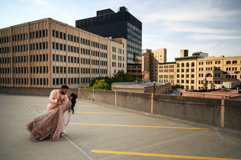 A couple dressed in traditional wedding attire shares a romantic dance on a rooftop parking lot with a cityscape of tall buildings in the background. The groom, wearing a light-toned outfit, dips the bride, who is wearing an ornate pink gown, under a twilight sky.