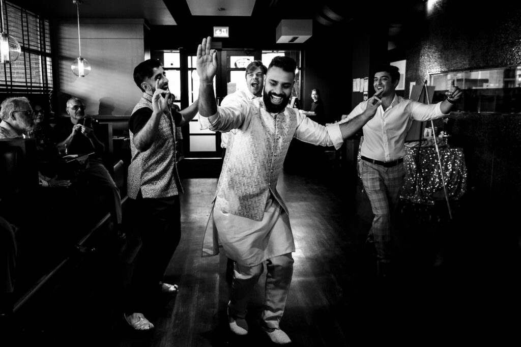 A group of men dressed in festive attire joyfully dance together in a dimly lit room. The central figure, wearing a traditional outfit, leads with energetic moves while others follow, smiling and clapping. Seated people in the background watch and applaud their performance.