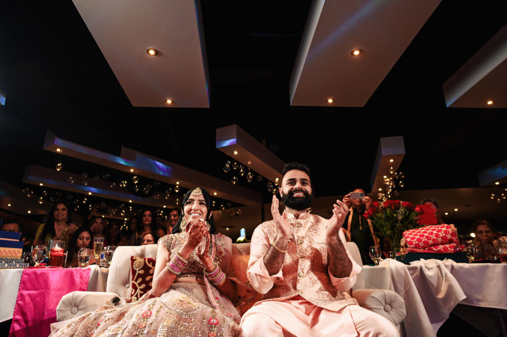 A couple dressed in traditional attire sits on a white couch and claps, smiling and looking ahead. The woman wears an ornate lehenga while the man dons a peach-colored kurta. They are surrounded by guests and festive decor, with presents placed on a table nearby.