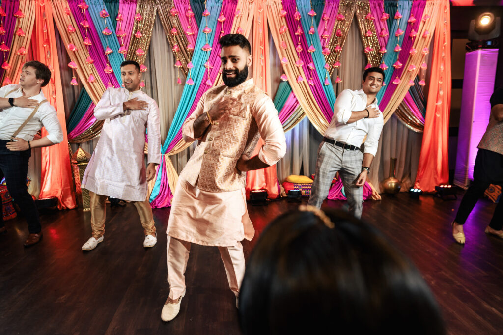 A group of five men performing a dance routine in front of vibrant and colorful drapery with hanging floral decorations. They are all smiling and wearing traditional attire, with the central dancer in a peach kurta and beige pants leading the synchronized movements.