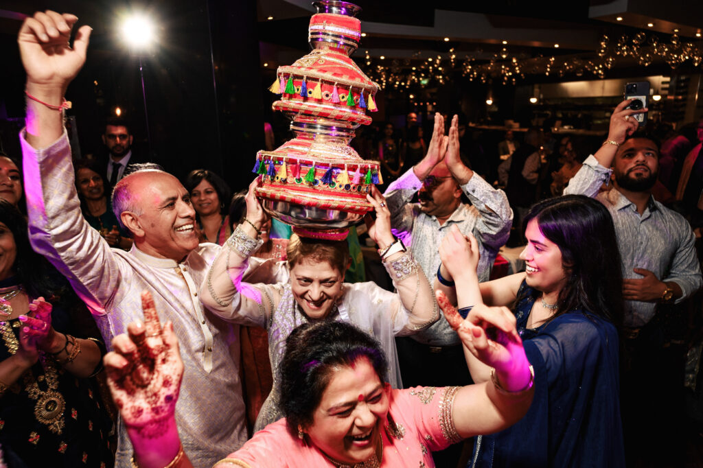 People celebrating at a festive event. A person in the center is smiling and balancing a colorful, decorated pot on their head while others around them cheer and dance. The atmosphere is lively, with many people dressed in traditional attire and joyful expressions.