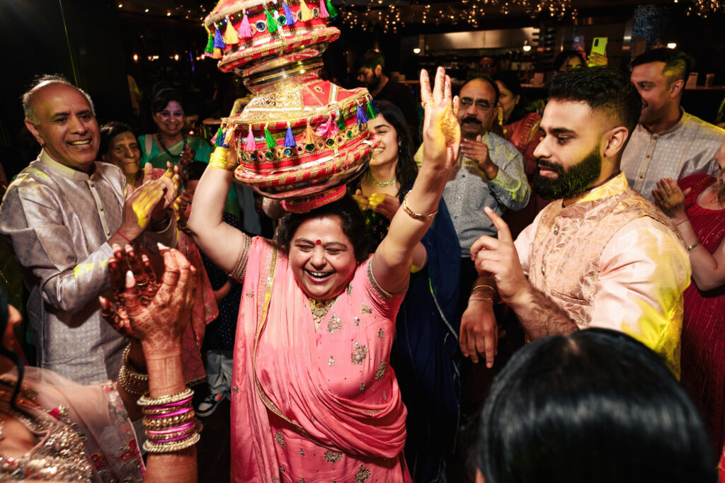 A woman dressed in a pink outfit and smiling, balances a colorful, traditional pot on her head while others around her cheer and clap, celebrating an event. The background is adorned with string lights, and everyone appears joyful and engaged in the celebration.