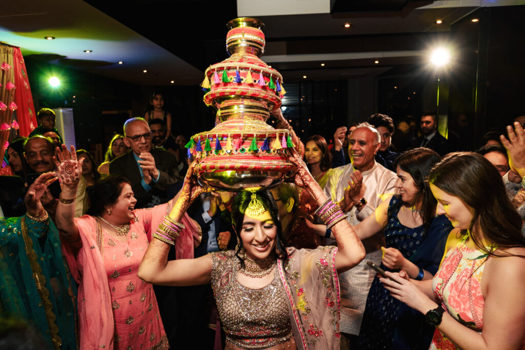 A woman dressed in traditional attire carries a large decorated vessel on her head at a lively celebration. She is surrounded by smiling people clapping and dancing, adorned in colorful clothing, under bright lights, creating a festive atmosphere.