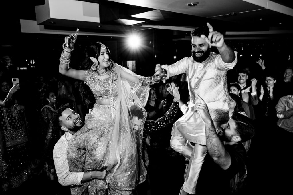 A joyful bride and groom, dressed in traditional Indian attire, are lifted on the shoulders of their friends during a lively celebration. Both are raising a hand in excitement, surrounded by cheering guests in a festive, illuminated indoor setting.