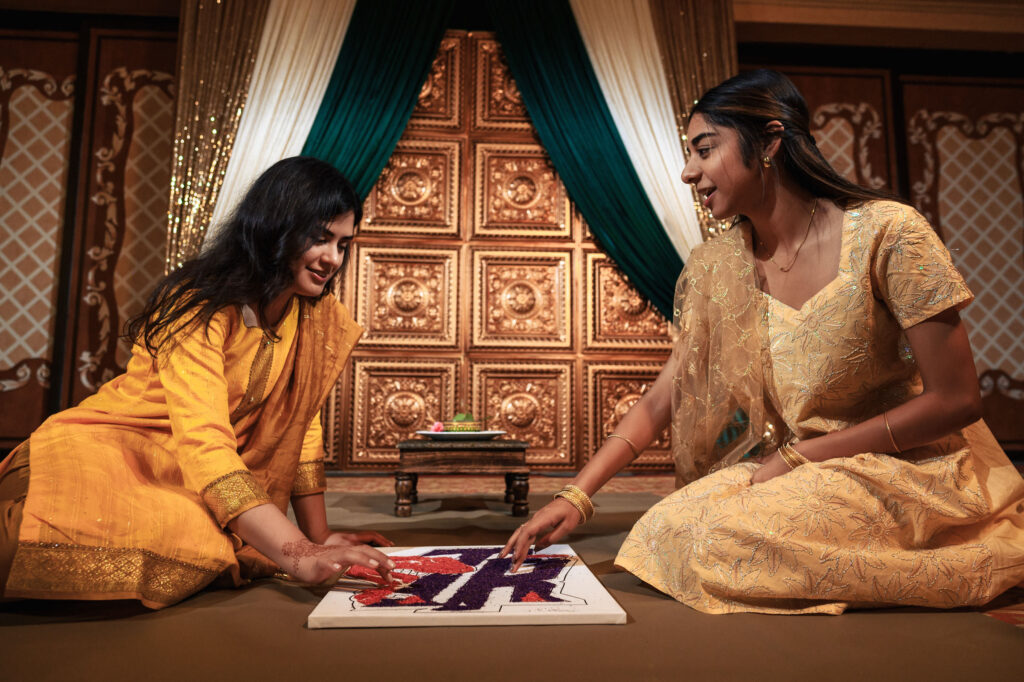 Two women in traditional attire are engaging in creating a colorful, intricate design on the floor using colored powders. They are seated on a richly decorated carpet, with an ornate wooden backdrop and green curtains behind them.