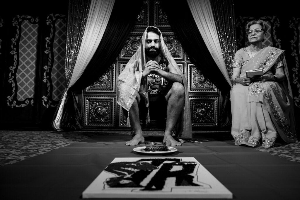 A man sits cross-legged, wearing a shawl over his head, with an older woman beside him. Both are dressed in traditional attire. In front, there's a canvas with the letters "SR" painted boldly. The background features ornate, decorative panels and curtains.