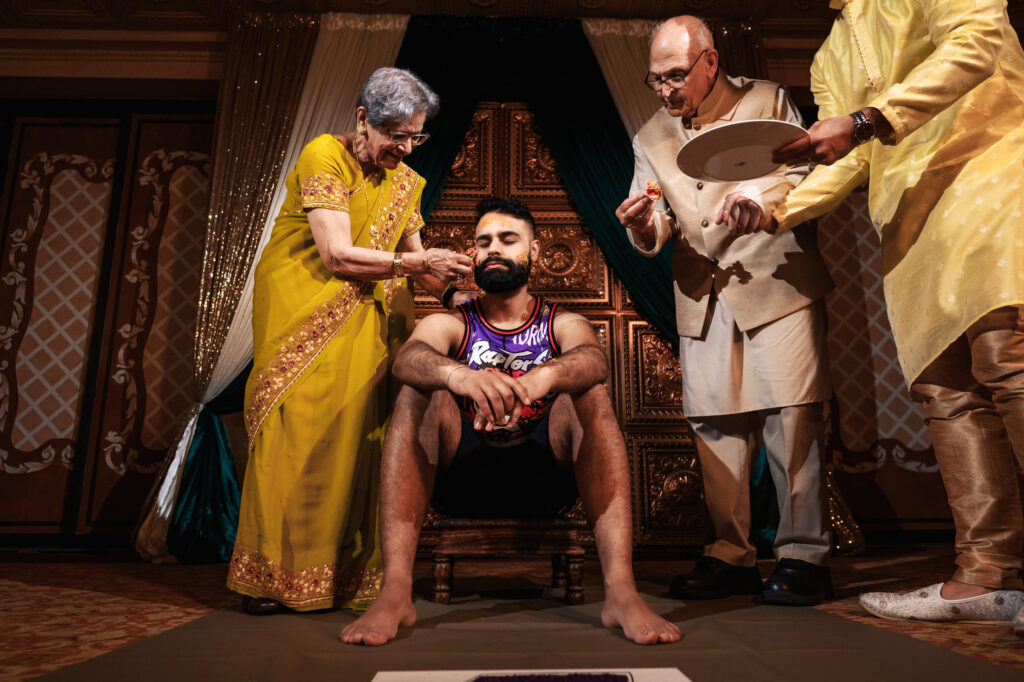 A man sitting on a decorated chair is being fed by an elderly woman in a yellow traditional outfit, while an elderly man in formal attire assists. Another person in traditional clothing holds a plate nearby. The setting is a richly decorated room with intricate carvings.