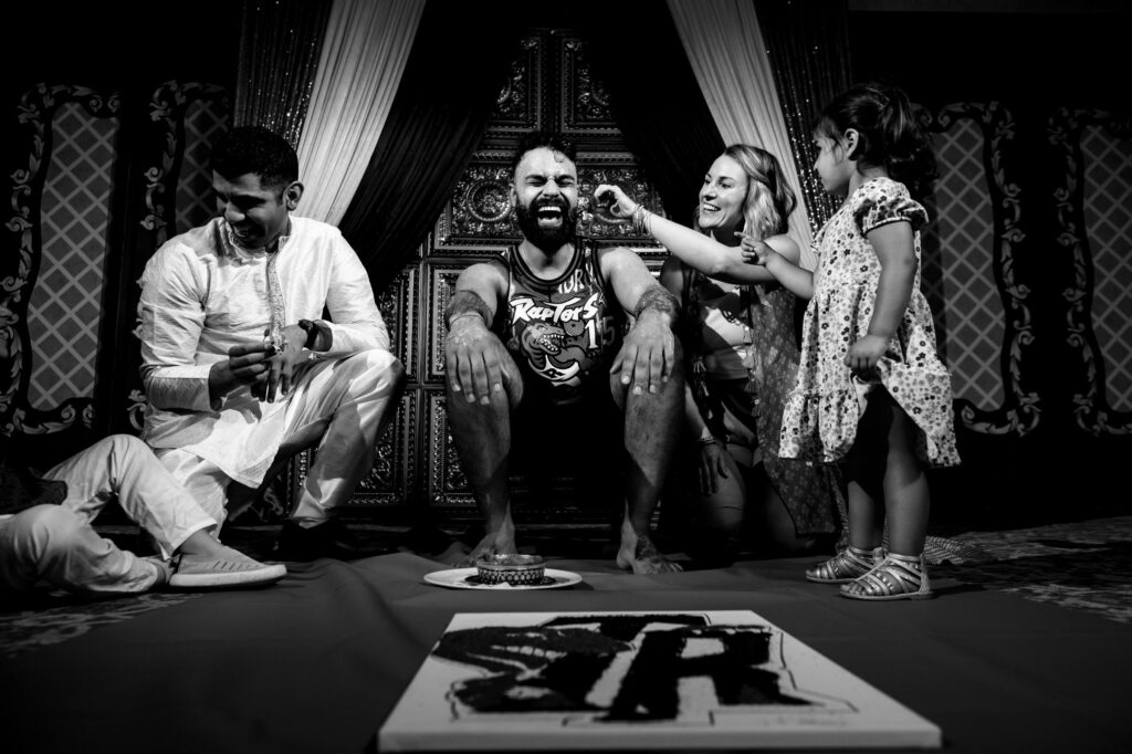 A man laughs while a woman and a child playfully smear something on his face. Another man dressed in traditional attire sits next to them, also smiling. They are all seated on a decorated stage, with a painting on the floor in front of them. The scene is joyful.