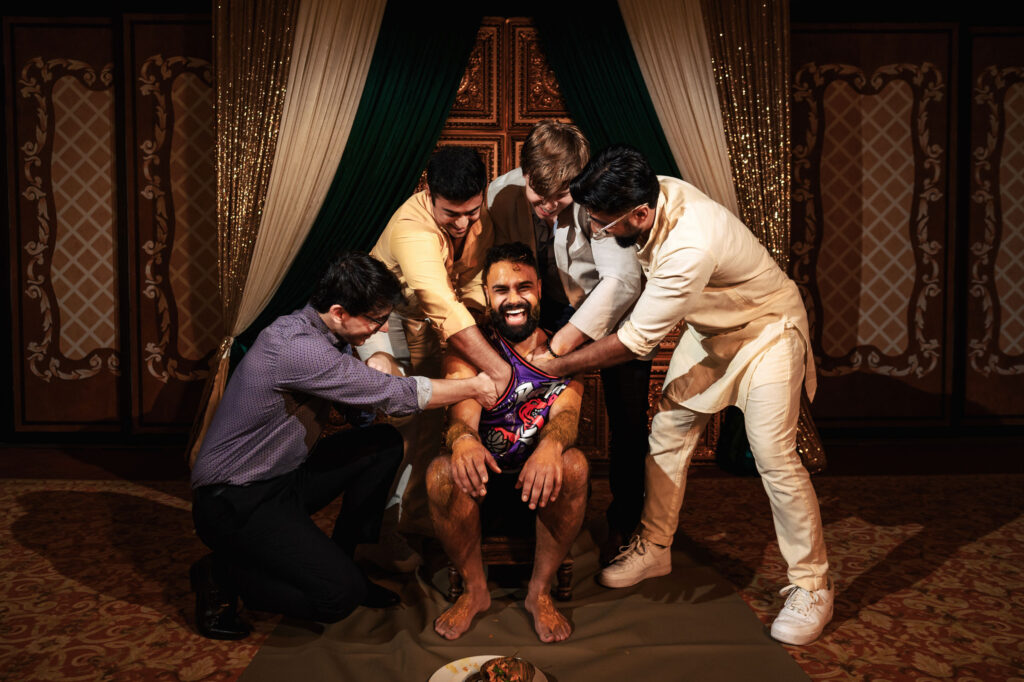 A joyful man sits on a chair, wearing colorful clothing, surrounded by four smiling men who are applying turmeric paste to his body. The background features ornate golden curtains and a decorative screen. A plate of traditional items is on the floor near him.