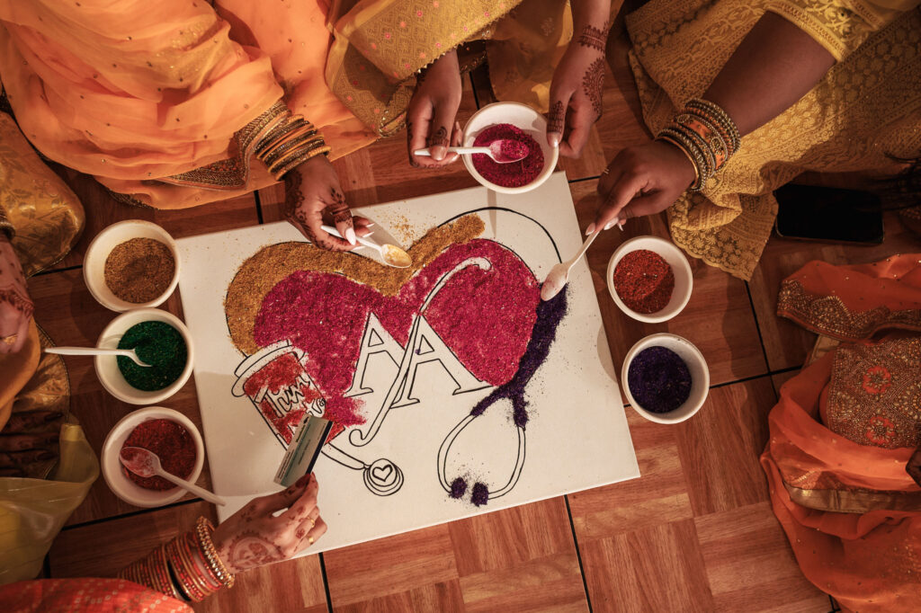 A group of people in vibrant traditional attire create an intricate rangoli design with various colored powders on a floor. The design features two intertwined hearts and the letter "A" in the center. The scene is full of festive and collaborative energy.