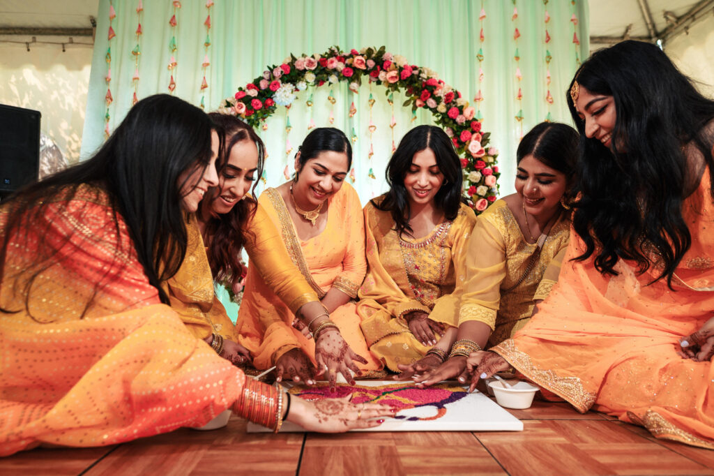 A group of women in vibrant yellow and orange traditional attire sit on the floor, engaged in creating a colorful rangoli design. They are surrounded by festive decorations, including a floral garland arch in the background, and they appear joyful and focused.