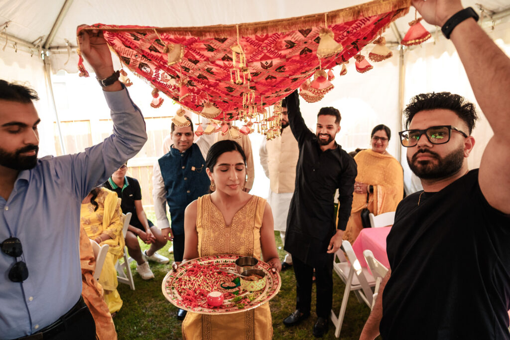 A woman in an ornate gold outfit walks under a decorative canopy held by four men, while carrying a tray with flowers and a candle. Other people, dressed in traditional attire, are seated and standing under a tent in the background.