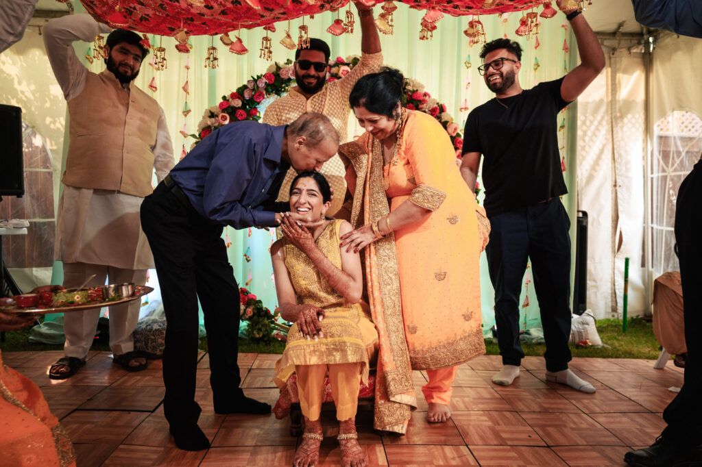 A joyful scene at a pre-wedding ceremony. A smiling woman in a yellow dress sits as an older man kisses her forehead and a woman placed a hand on her shoulder. Observers dressed in traditional attire watch the moment. The setting is decorated with flowers and drapes.
