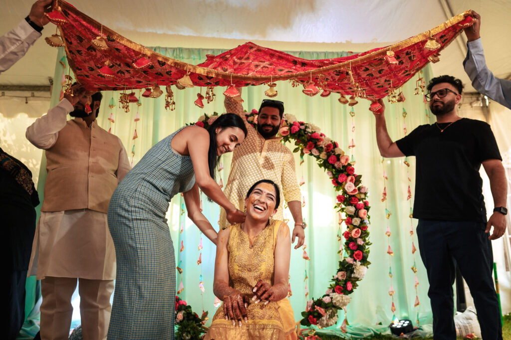 A woman, dressed in a yellow outfit, is seated and smiling joyfully under a decorative red canopy. Another woman in a blue outfit is applying henna to her arm. Two men hold up the canopy. The background features a floral arch with red, white, and green decorations.